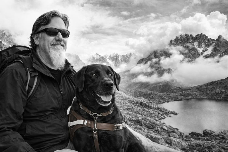 Ted and his guide dog fauna sit on top of a mountain. In the background a lake and mountian with clouds can be seen.